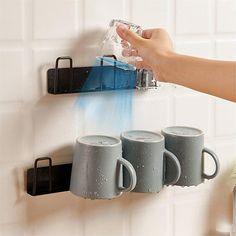 three mugs are hanging on the wall with water coming out of them and being held by someone's hand