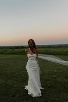 a woman in a white dress is standing on the grass with her veil blowing in the wind