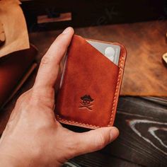 a hand holding a brown leather card case on top of a wooden table next to a wallet