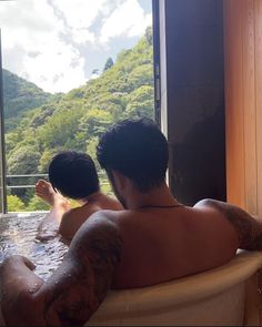 two men sitting in a bathtub looking out the window at trees and mountains behind them