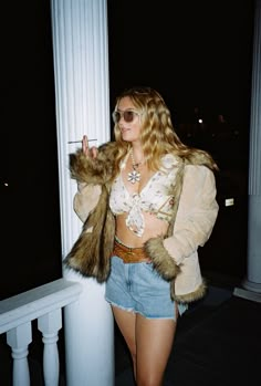 a woman in short shorts and a fur coat standing on a porch with her hand up to her mouth