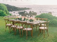 a table set up with flowers and place settings on the grass by the water at sunset