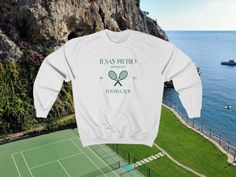 a white sweatshirt with a tennis racket in front of the ocean and a boat