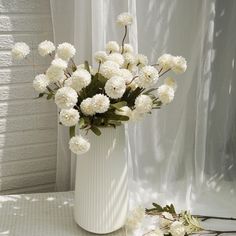 a white vase filled with lots of white flowers next to a window covered in curtains