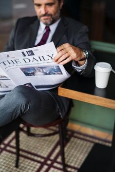 a man is sitting at a table reading the daily newspaper while holding a coffee cup