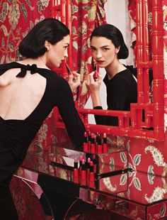 two women in front of a mirror with lipstick on the table and one holding a brush