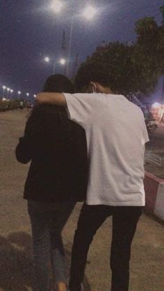 two people are walking down the street at night with their arms around each other's shoulders