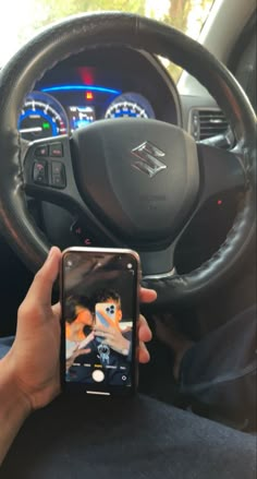 a person holding up a cell phone in front of a car steering wheel and dashboard