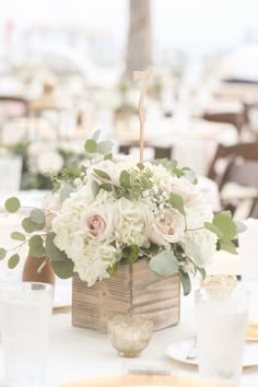 an arrangement of flowers in a wooden box on a table with white linens and place settings