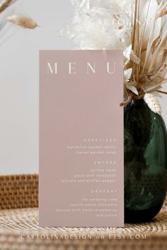 a pink menu card sitting on top of a green vase filled with white flowers and feathers