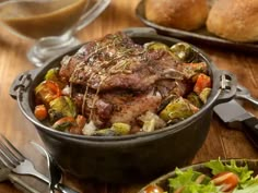 a pot filled with meat and vegetables on top of a wooden table next to silverware