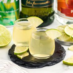 two glasses filled with white wine and limes on a table next to alcohol bottles