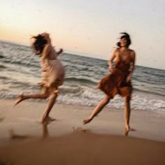 two women are running on the beach near the ocean at sunset or sunrise, one is wearing a brown dress and the other is in white