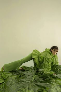 a woman in green is laying on the ground surrounded by plants and leaves with her legs spread out