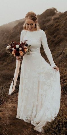 a woman in a long white dress holding a bouquet on top of a hill near the ocean