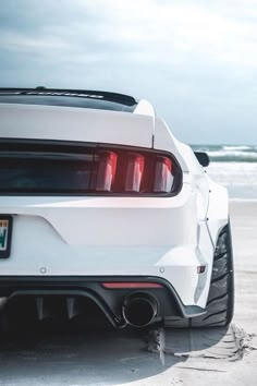 the rear end of a white sports car parked in front of the ocean on a beach