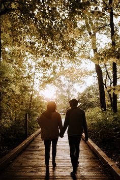 two people holding hands walking down a wooden walkway in the woods with trees on either side