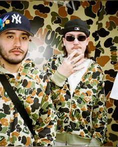 two young men standing next to each other in front of a wall covered with camouflage