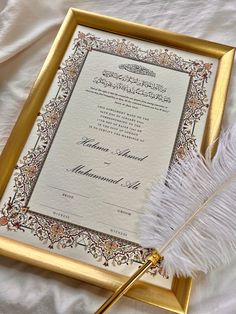 an ornate wedding card and feather on a white bed sheet with gold trimmings