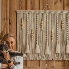 a woman holding a cat in front of a wall hanging with tassels on it