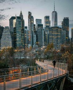 a person walking across a bridge in front of the city skyline at dusk with lights on