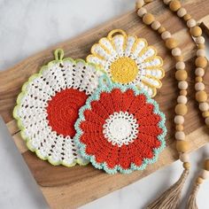 two crocheted flower coasters sitting on top of a wooden board next to a beaded necklace