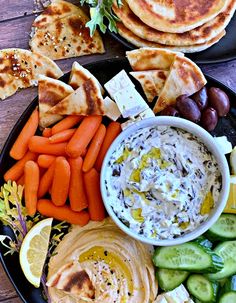 a black plate topped with hummus, carrots, cucumbers and crackers