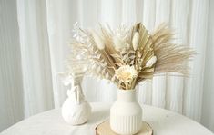 two white vases with dried flowers in them on top of a round tablecloth