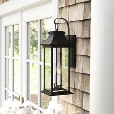 a black lantern hanging on the side of a house next to white flowers and an open window