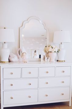 a white dresser topped with lots of drawers next to a wall mounted mirror and two lamps