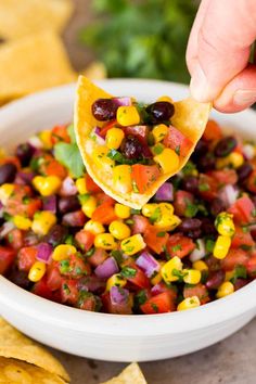 a tortilla chip being held over a bowl filled with corn and black bean salsa