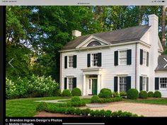 a large white house with black shutters on the front and side windows, surrounded by lush green trees