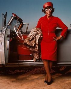 a woman in a red dress and leopard print scarf leaning on the back of a car