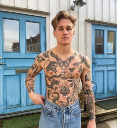 a man with tattoos standing in front of a blue door wearing jeans and holding his hands on his hips