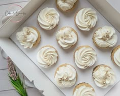 twelve cupcakes with white frosting in a box on a wooden table next to flowers