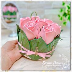 a hand holding a small pink rose in a paper basket with leaves and beads on it