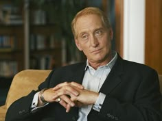 a man in a suit sitting on a couch with his hands folded and looking at the camera