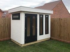 a small white shed sitting in the grass next to a wooden fence and brown brick building