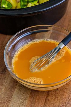 a glass bowl filled with orange liquid next to a metal whisk on top of a wooden table