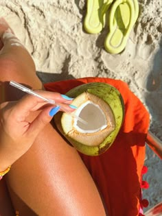 a woman sitting on the beach with a coconut in her hand and a straw in her other hand