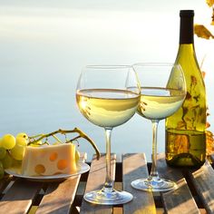 two glasses of white wine and cheese on a wooden table with water in the background