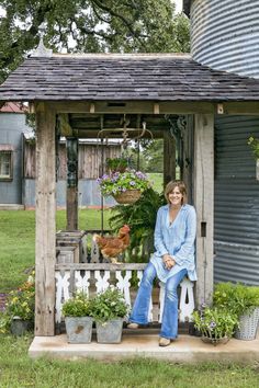 a woman sitting on top of a wooden bench in front of a shed filled with potted plants