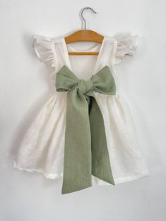 a white dress with a green bow hanging on a wooden hanger against a wall