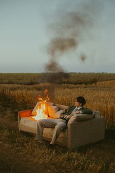 a man sitting on top of a couch next to a fire in the middle of a field