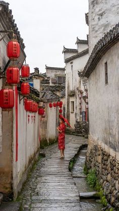 a woman in a red dress walking down an alley way with lanterns hanging from the buildings