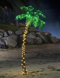 a lighted palm tree in the middle of a beach with rocks and water behind it