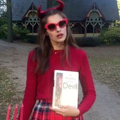 a woman wearing red glasses holding a book in front of a house with devil horns on her head