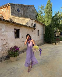 a woman in a purple dress is holding flowers and talking on her cell phone while standing outside an old building