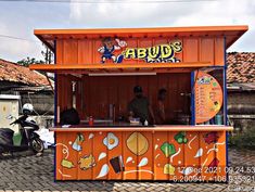 an orange and blue food stand on the side of a road with people standing around it