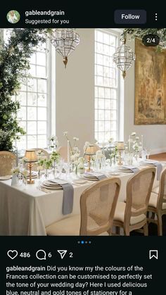 the table is set with white flowers and place settings on it, along with an arrangement of greenery